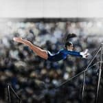 A female gymnast in mid air reaching for the parallel bars.